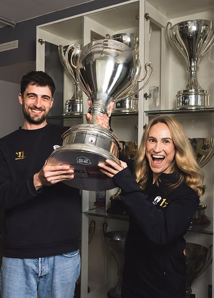 Two people smiling and holding a large silver trophy in front of a cabinet filled with trophies.