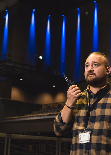 Man in plaid shirt holding a walkie-talkie with blue stage lights in a theater.