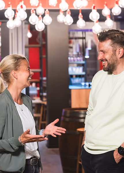 Smiling man and woman talking in a warmly lit indoor space with hanging lights overhead.