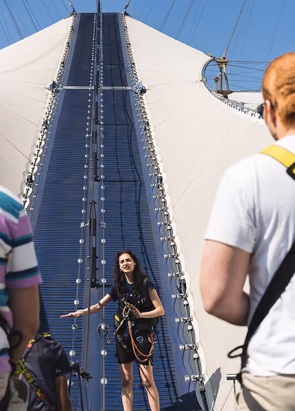 Woman in safety gear instructs people near a tall climbing wall on a sunny day.