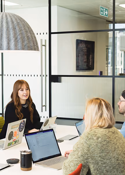 Three people with laptops engaged in a meeting at a modern office table under a large gray lamp.