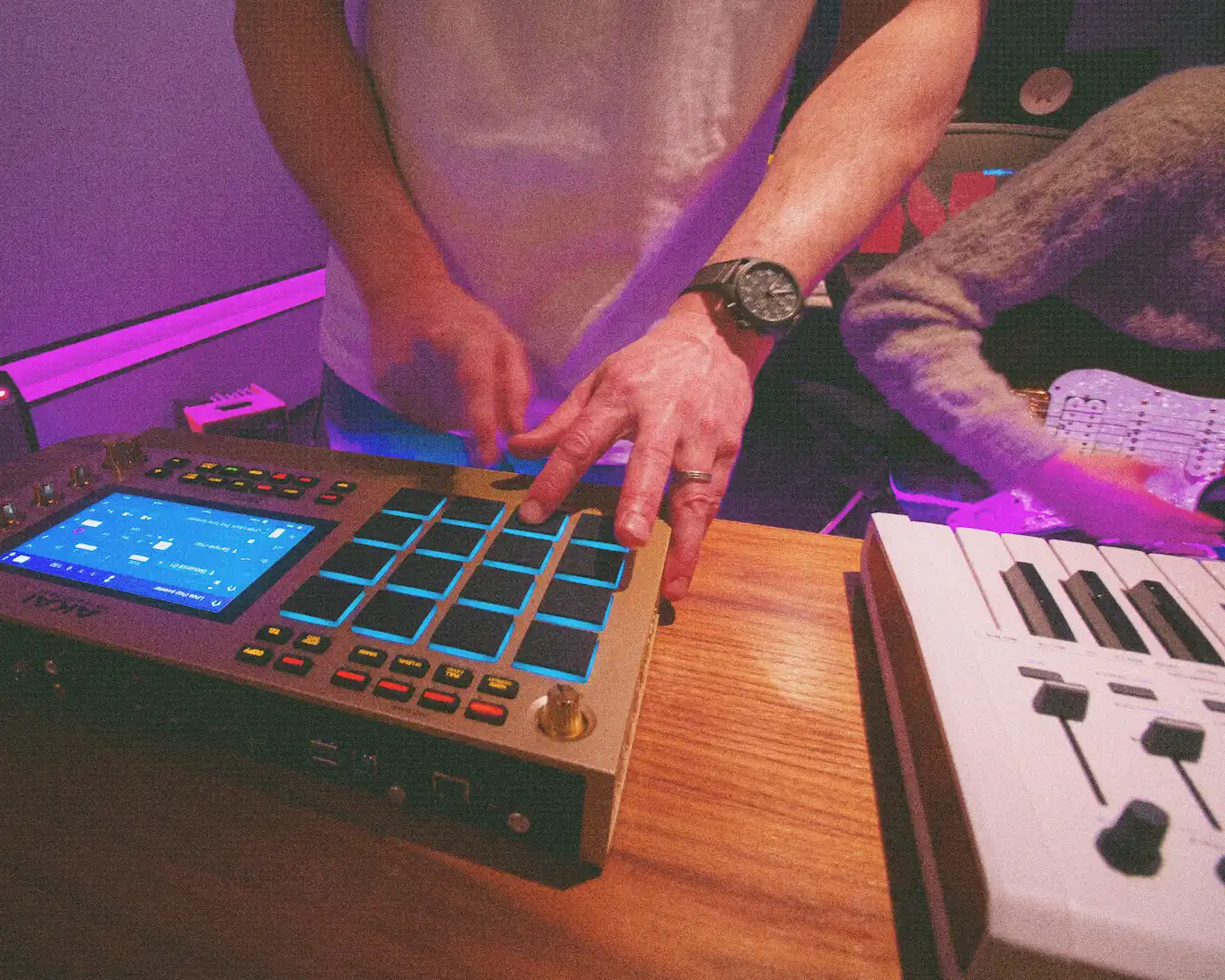 Person using a music production controller with illuminated pads and a second person playing a guitar next to a keyboard.