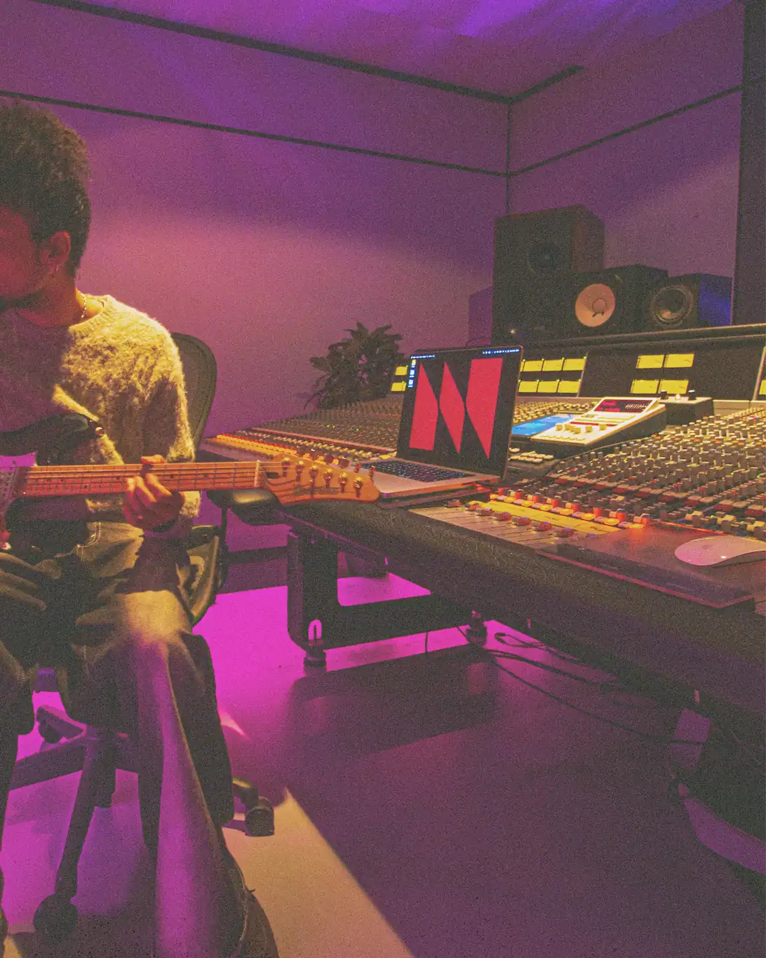 Person playing electric guitar in a purple-lit recording studio with mixing console and laptop.
