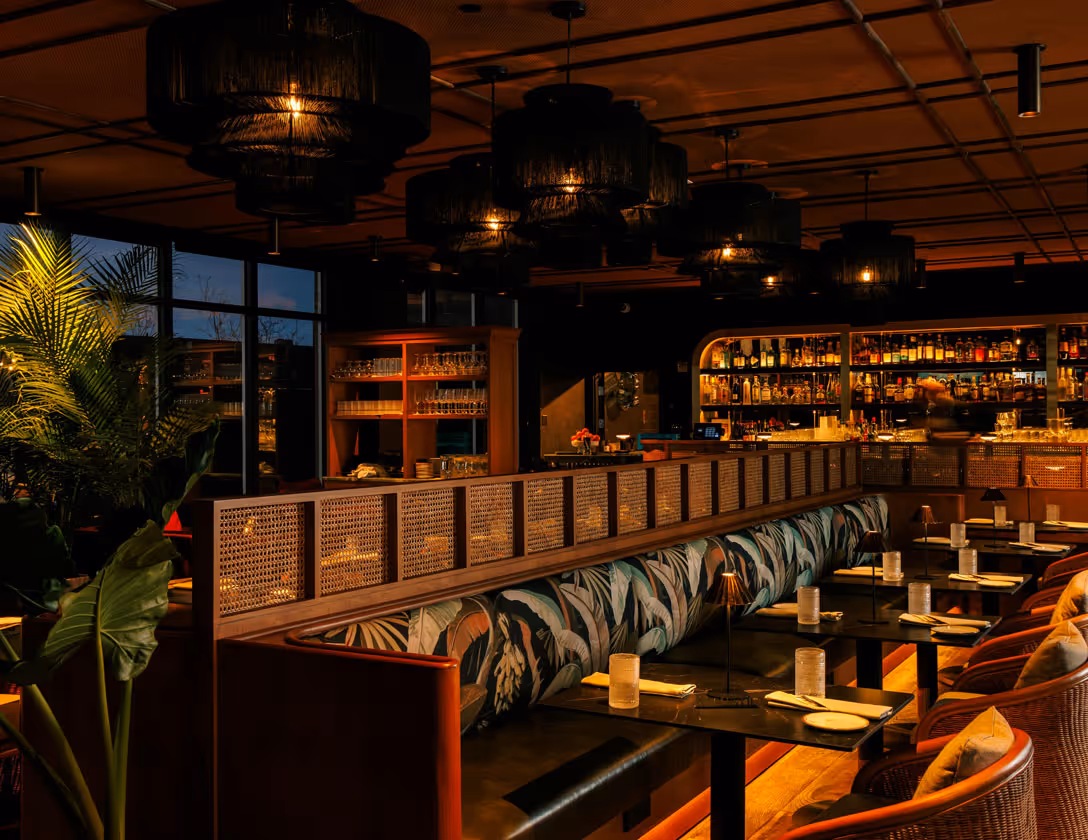 Cozy restaurant interior with patterned banquette seating, wooden tables set with plates and napkins, warm pendant lighting, and a stocked bar in the background.