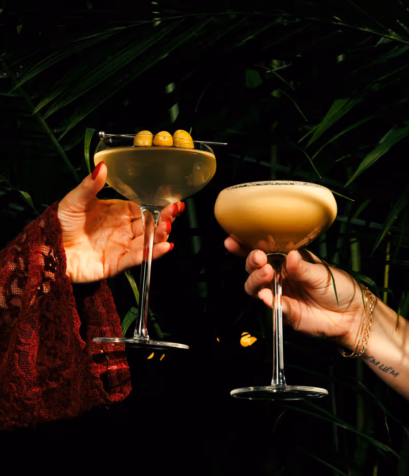 Two hands holding cocktails against a dark background of green palm leaves.