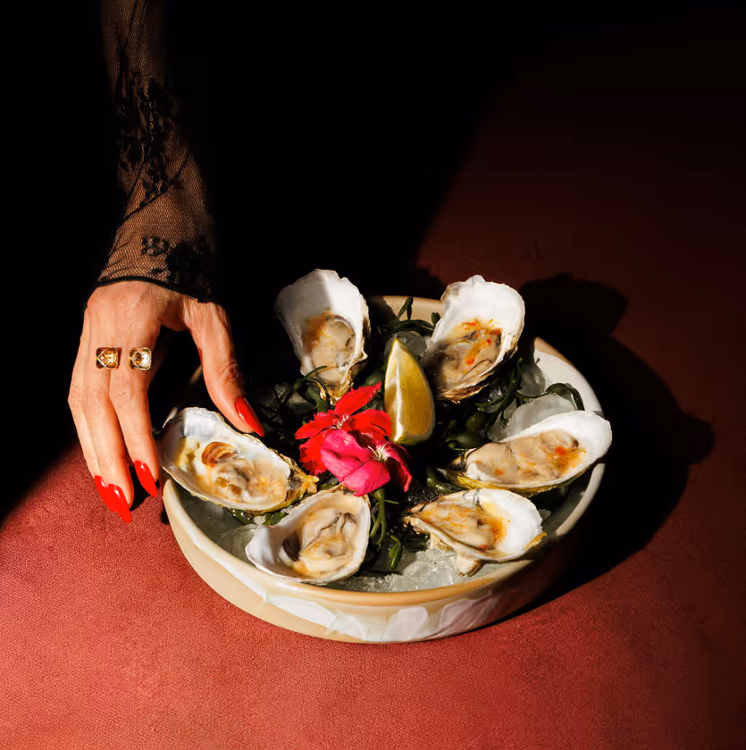 Six oysters on ice arranged in a bowl with a lemon wedge and a pink flower, a hand with red nails and rings reaching in