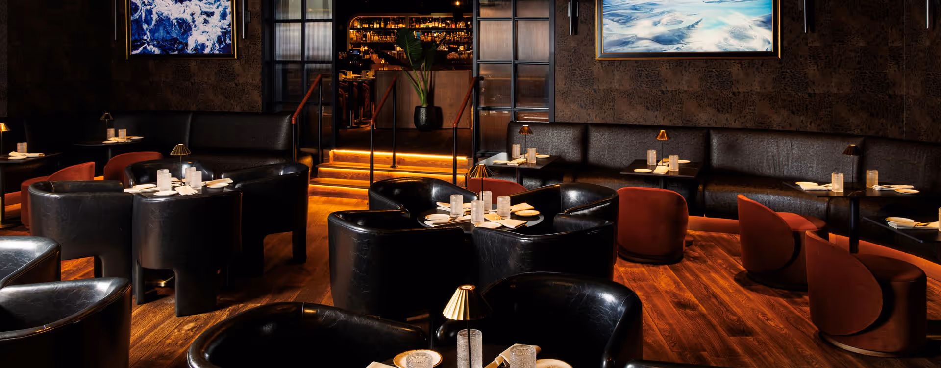 Luxurious restaurant interior with black leather chairs, red armchairs, dark wood floors, and ambient lighting leading to a bar area.