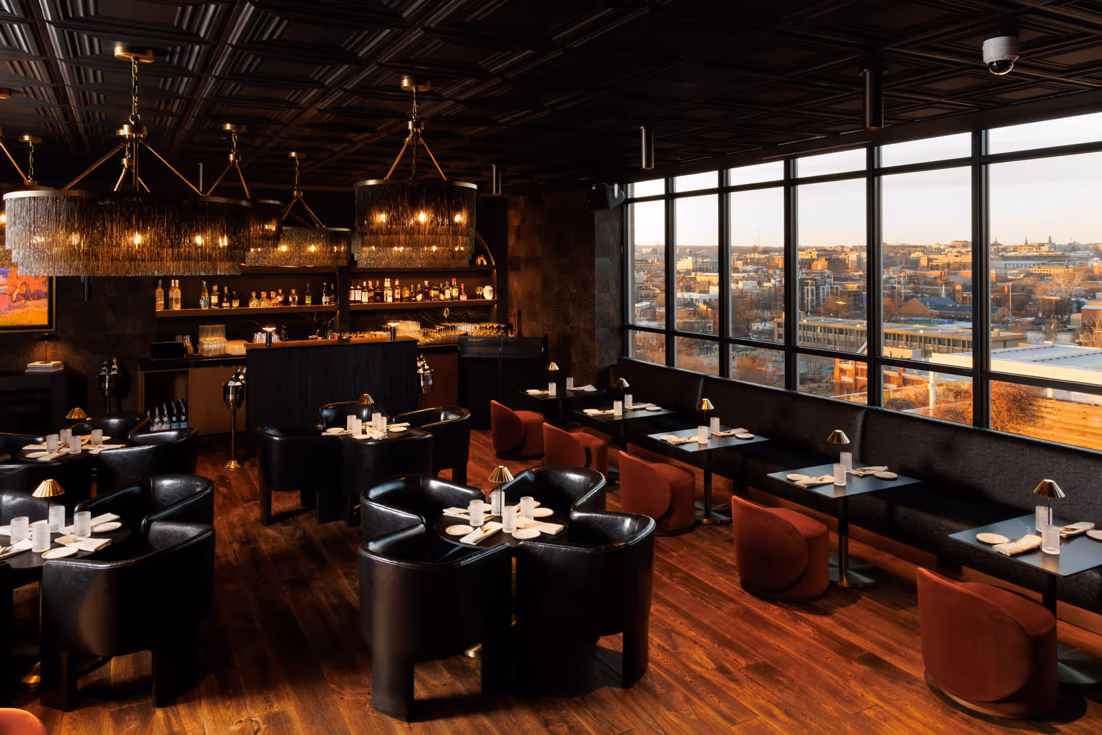 Elegant restaurant interior with black leather chairs, red velvet stools, dark wood flooring, modern chandeliers, and large windows overlooking a cityscape at sunset.