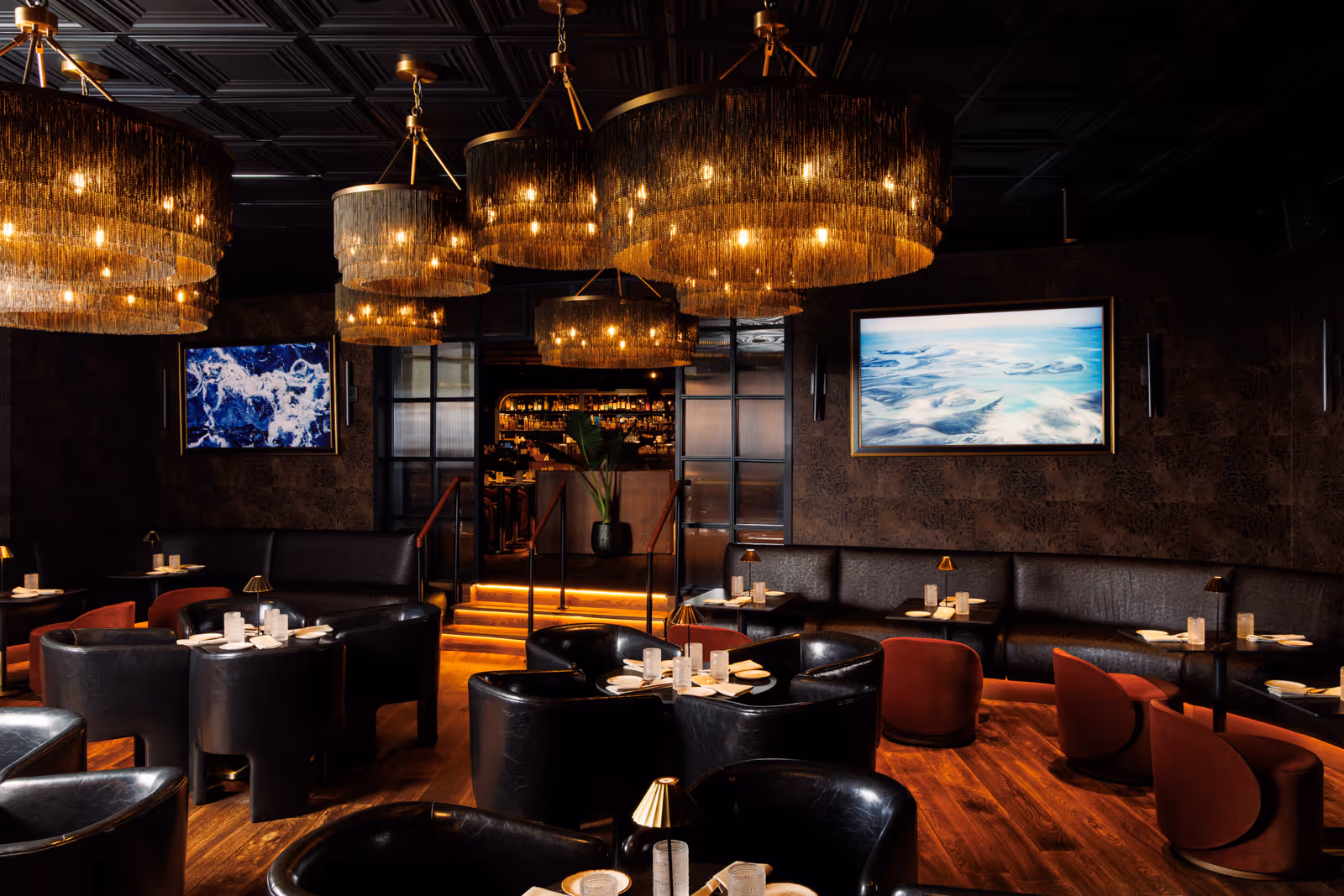 Elegant restaurant lounge with dim lighting, black leather chairs, red velvet stools, large chandeliers, and framed ocean artwork.