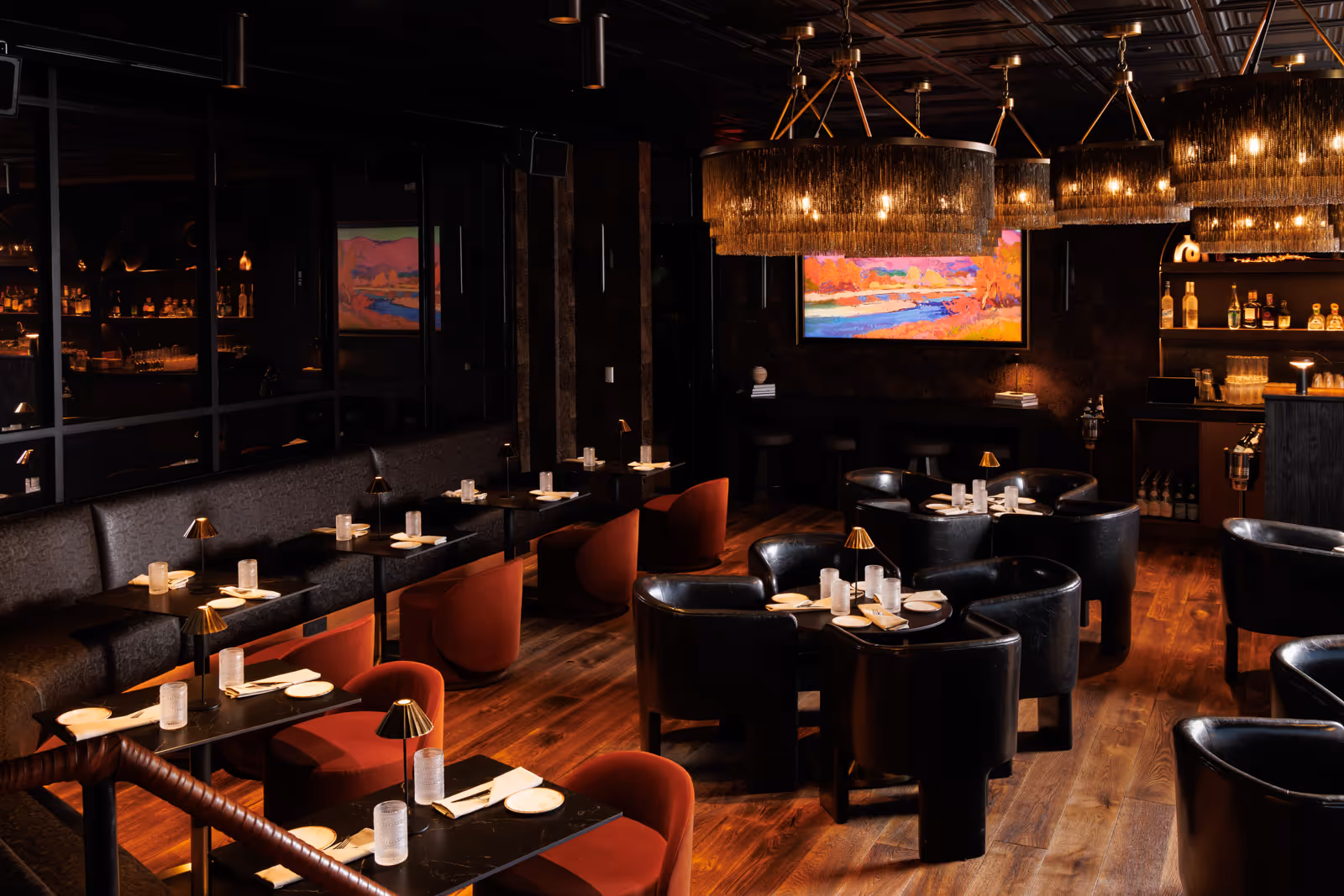 Dimly lit modern restaurant interior with black and red chairs, wooden floor, pendant chandeliers, and colorful landscape painting on the wall.
