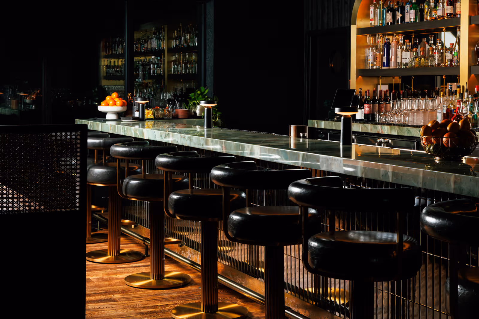 Dimly lit modern bar with black leather stools, a green marble countertop, bottles and glasses on shelves, and bowls of oranges on the bar.