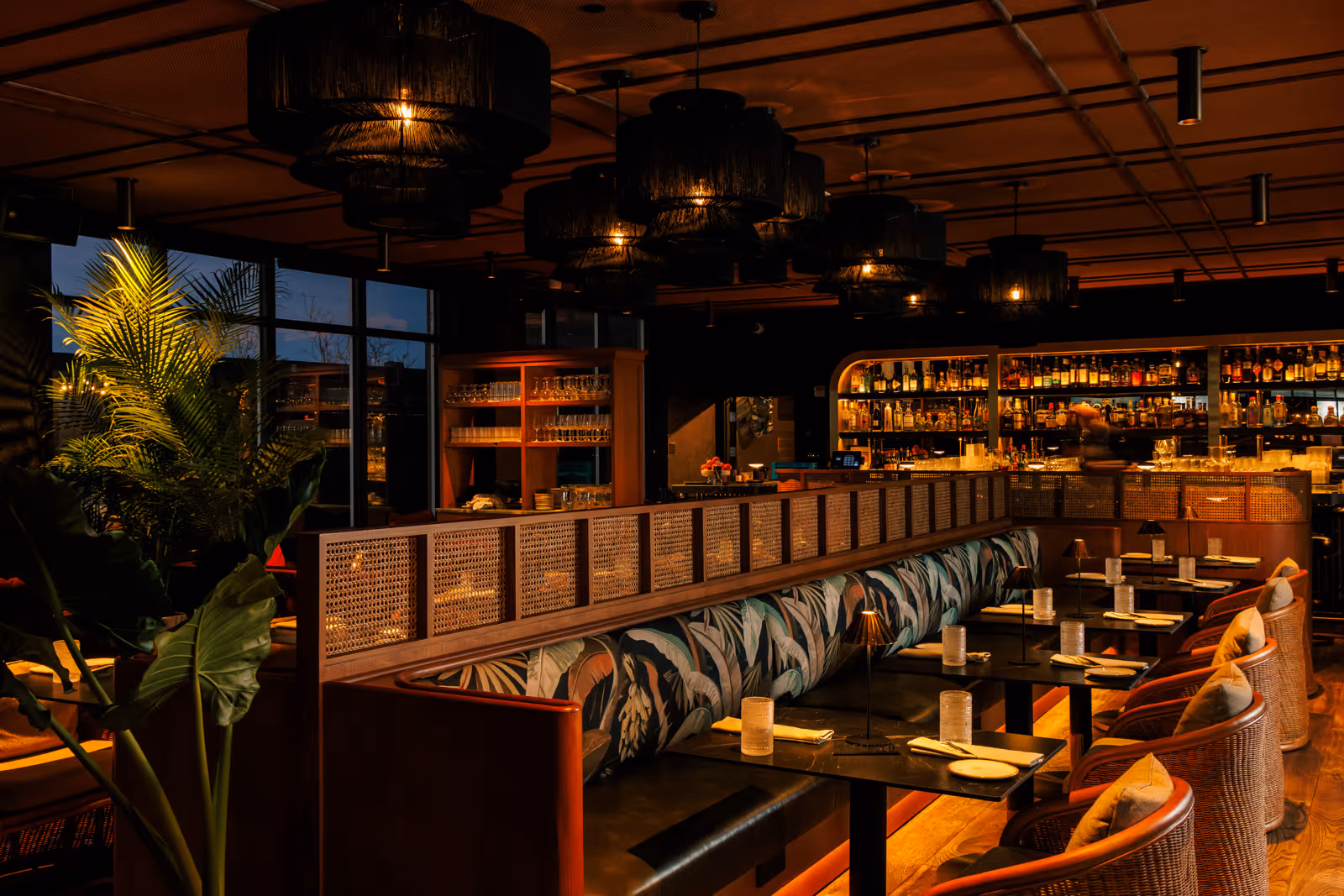 Cozy restaurant interior with patterned banquette seating, woven chairs, wood tables set with plates and glasses, large plants, and warm ambient lighting from multiple black pendant lamps.