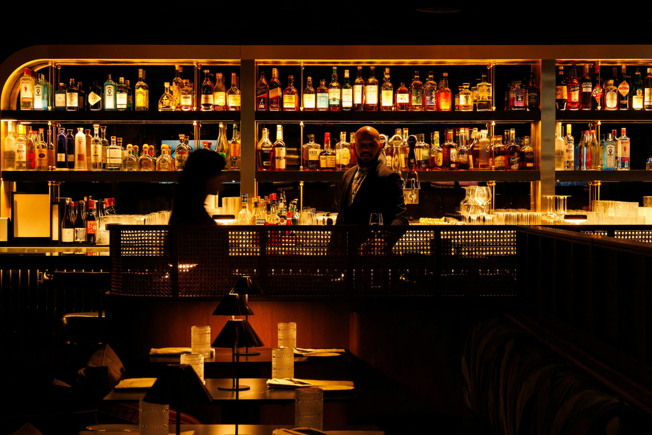 Dimly lit bar with shelves filled with liquor bottles. A bartender stands behind the counter, engaged with a customer. Cozy, intimate ambiance.