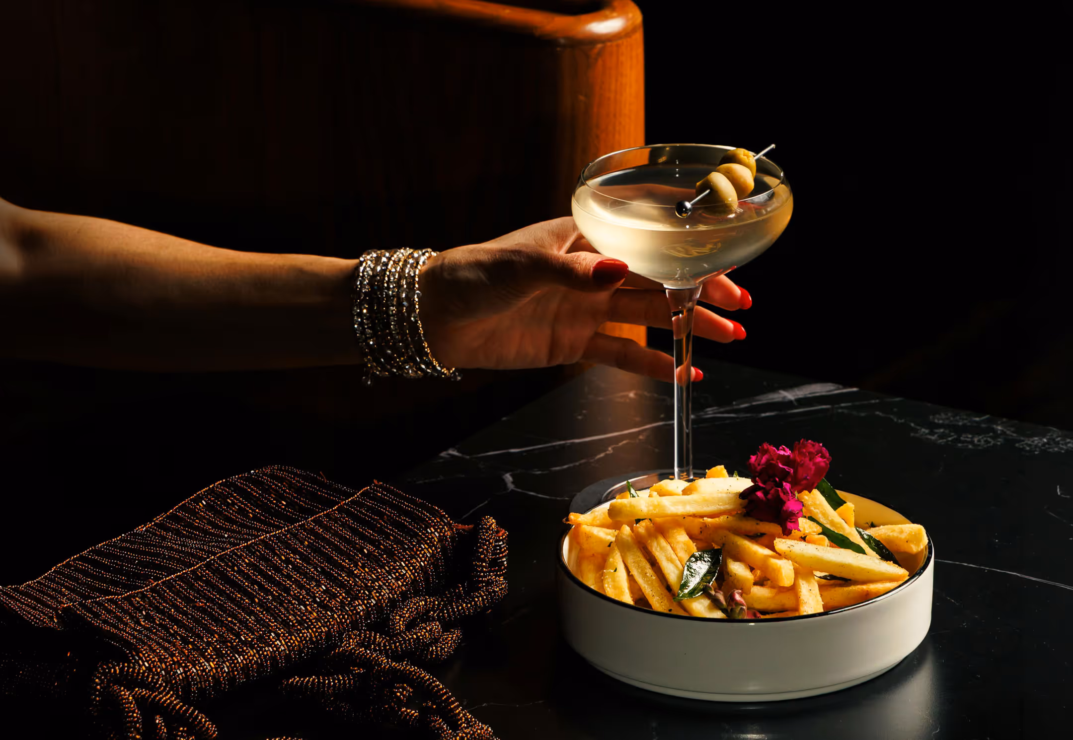 A hand adorned with bracelets holds a martini glass garnished with olives. Beside it, a bowl of golden fries with herbs and a flower sits on a dark table, conveying a luxurious, elegant atmosphere.