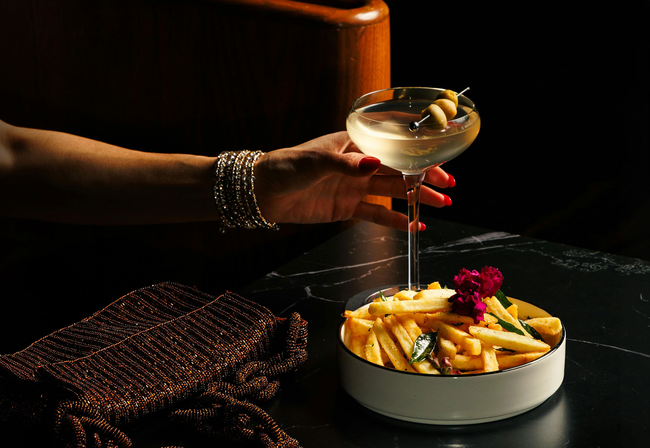 A hand adorned with bracelets holds a martini glass garnished with olives. Beside it, a bowl of golden fries with herbs and a flower sits on a dark table, conveying a luxurious, elegant atmosphere.