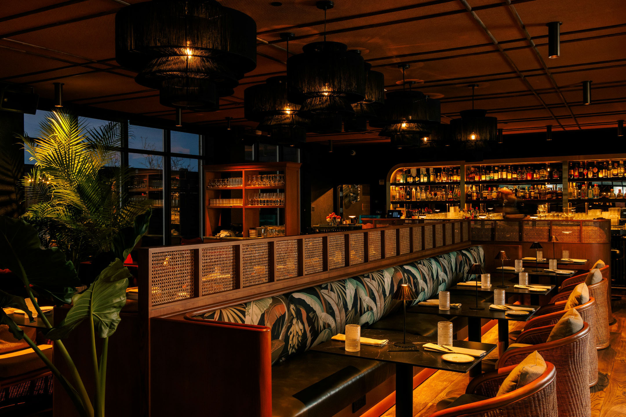 Chic restaurant interior with warm lighting, wooden furnishings, plush booths, and elegant decor. Open bar shelf with bottles in the background. Cozy ambiance.
