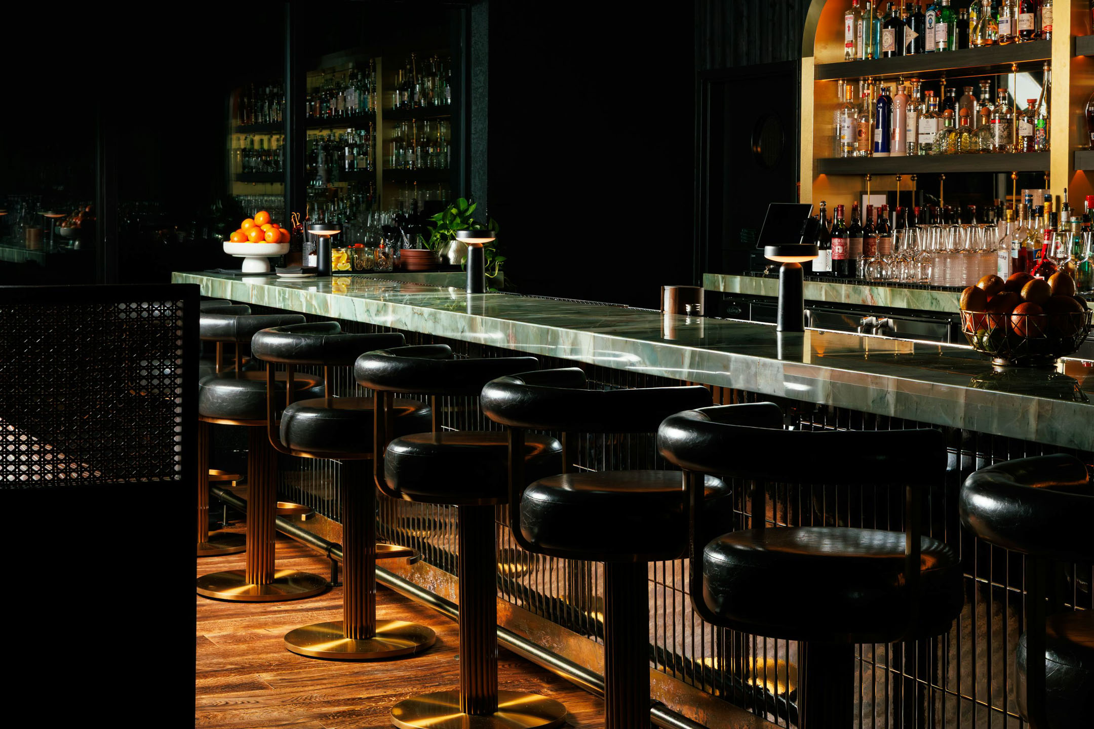 A dimly lit bar featuring a sleek marble counter lined with black stools. Shelves of bottles behind the counter and a bowl of oranges on the end of the counter.