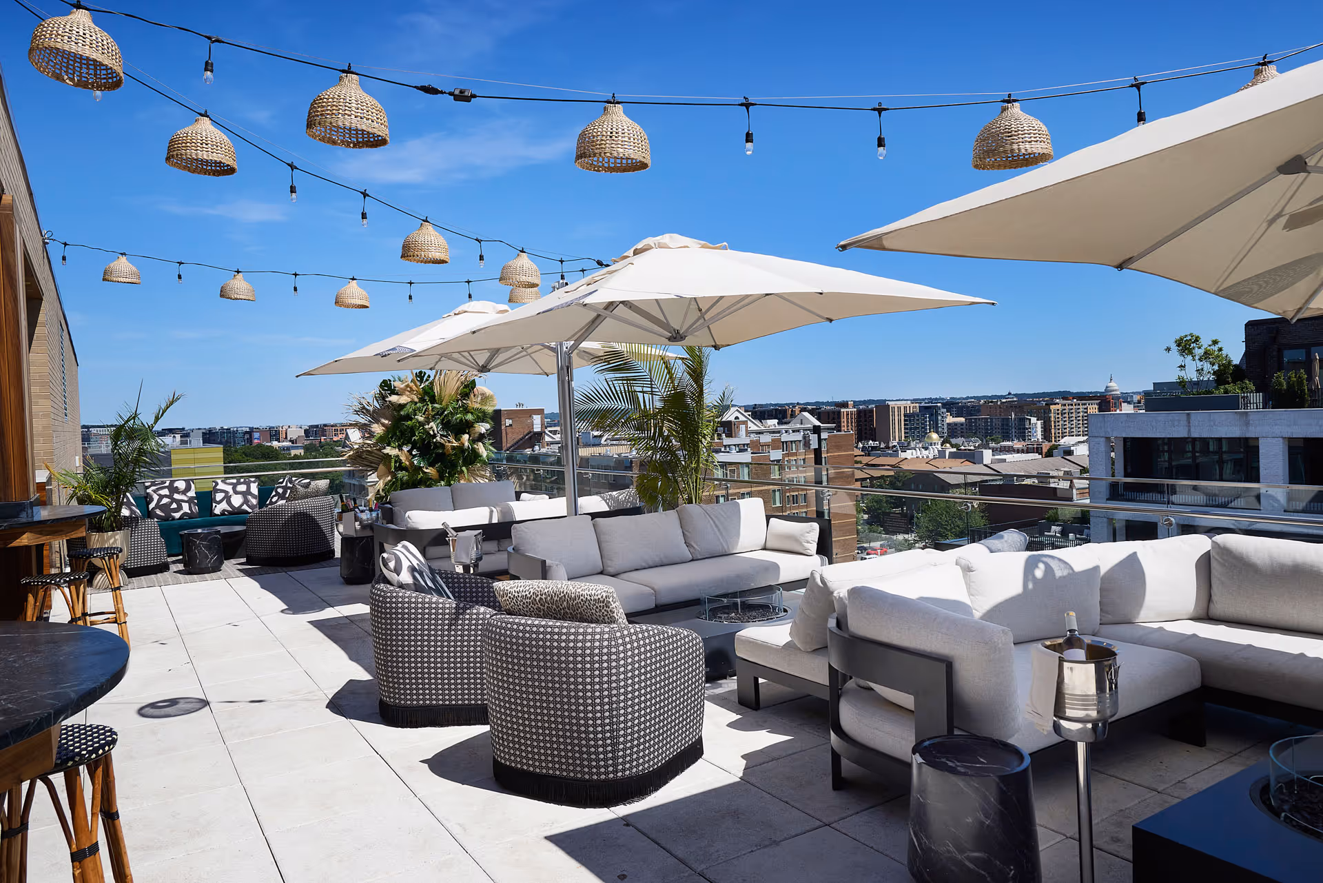 Daylight patio views of Washington DC at Realm Rooftop with tables and cream colored umbrellas and palms around the edge of the balcony and string lights above it all.