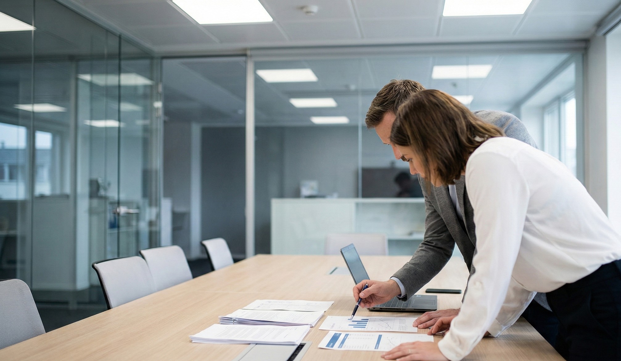 Two business professionals reviewing charts and documents on a conference table in a modern office.