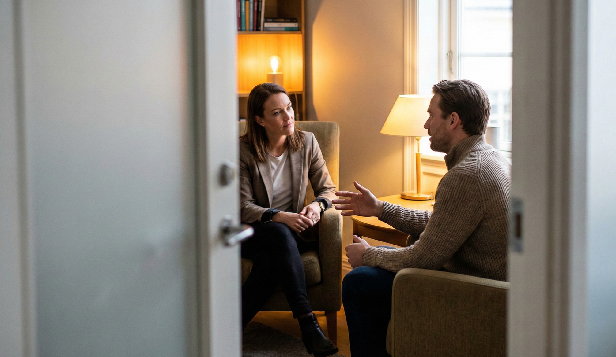 Two people having a serious conversation in a warmly lit room with armchairs and a table lamp.