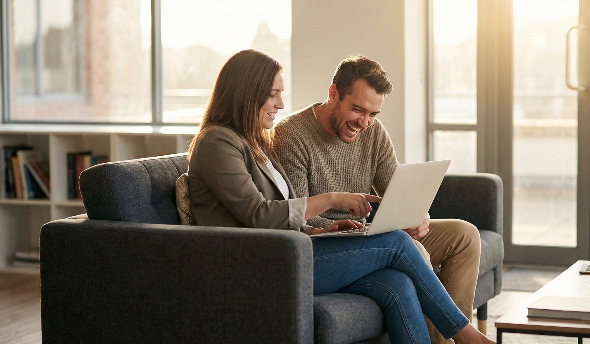 Smiling man and woman sitting on a gray couch looking at a laptop together in a sunlit room.