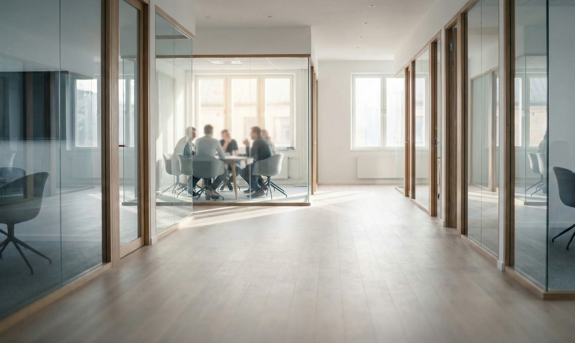 Office corridor with wood flooring and glass-walled meeting rooms, showing a group of people having a discussion in one room.