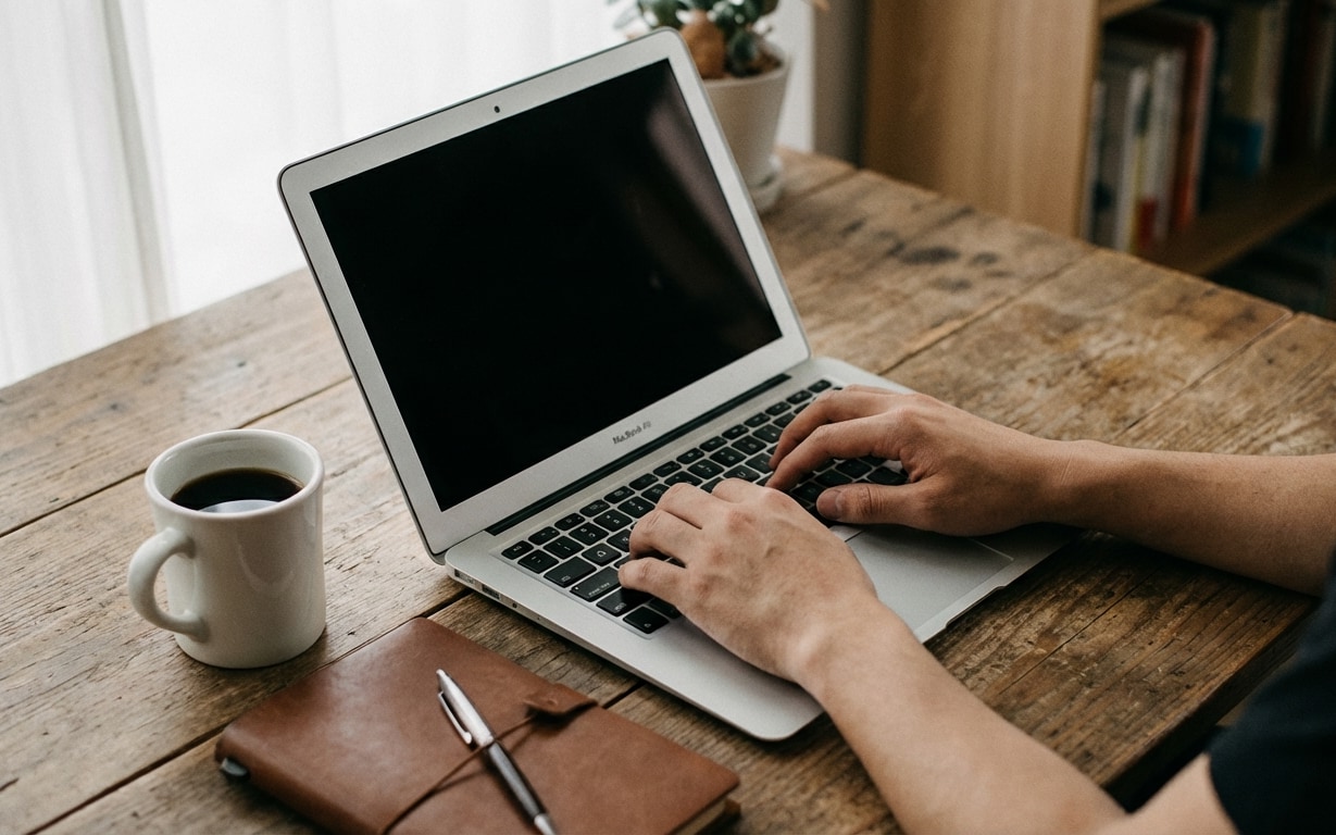 Person typing on a MacBook Air laptop on a wooden table with a cup of coffee and a brown notebook with a pen.