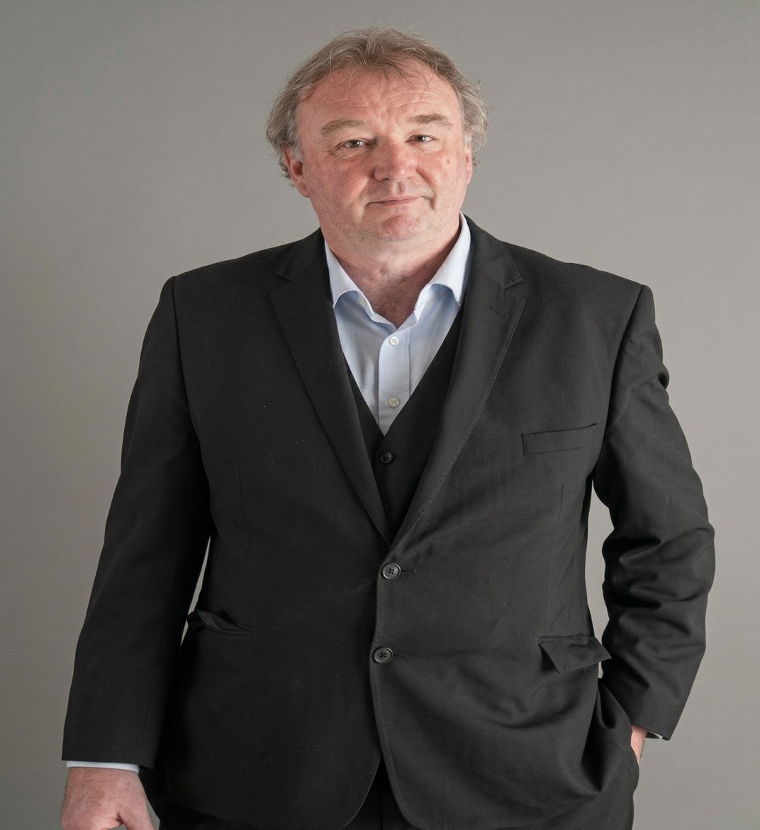 Middle-aged man with gray hair wearing a black suit and light blue shirt standing against a gray background.