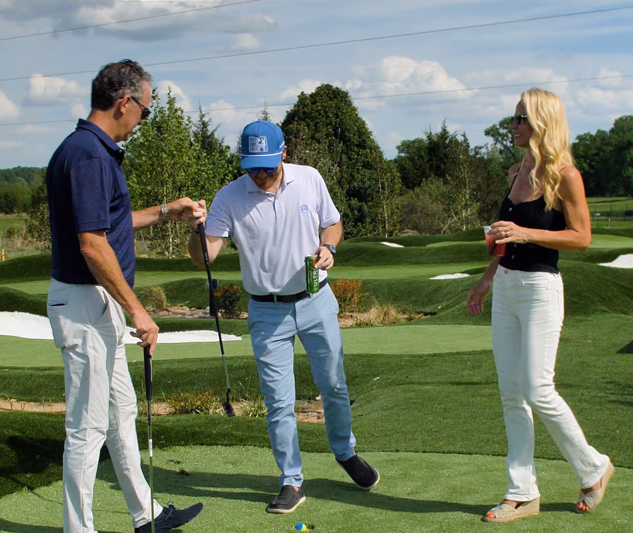 Two men fist bump on a golf course putting green while a woman nearby holds a drink and smiles.