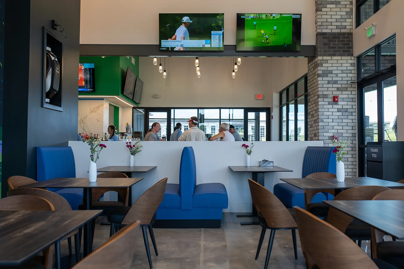 Outdoor evening view of a modern restaurant with large windows, string lights, umbrellas, and people mingling on a spacious patio.
