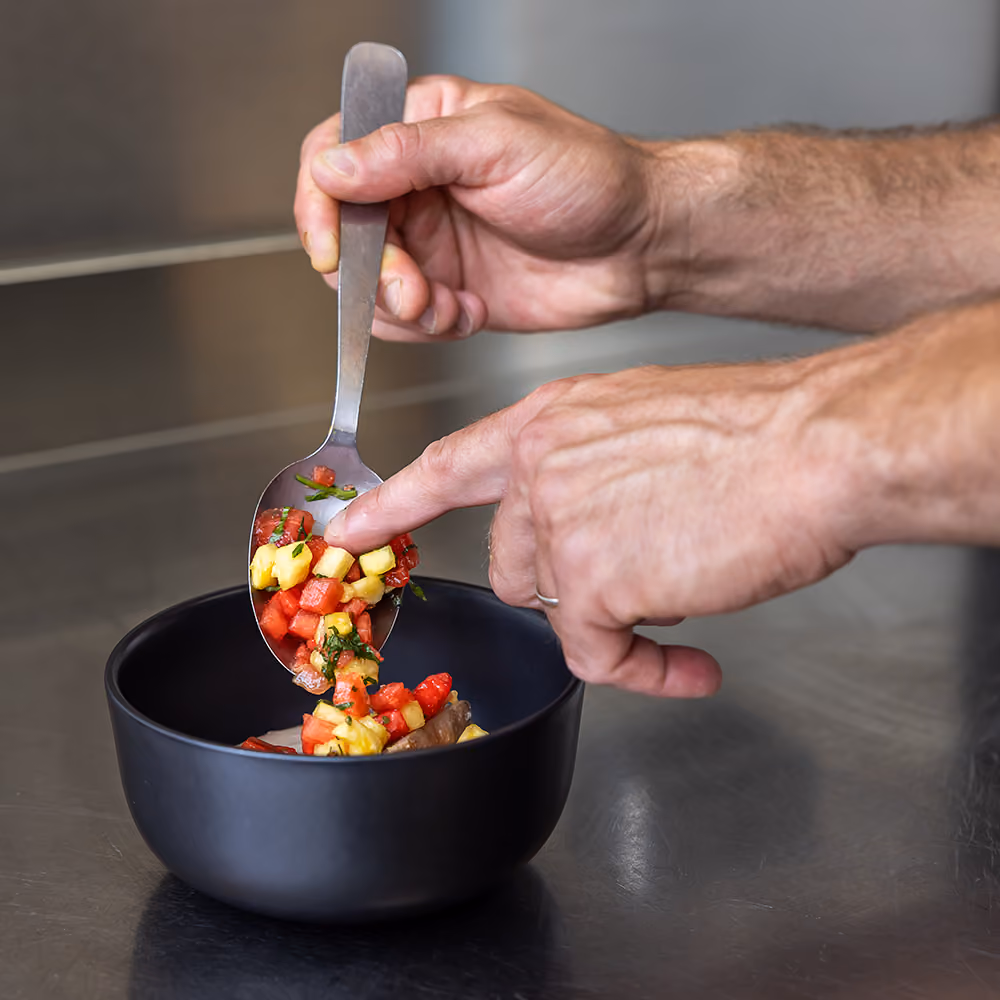 Chef dressant à la cuillère un tartare de pastèque, tomate et ananas dans un bol noir en cuisine