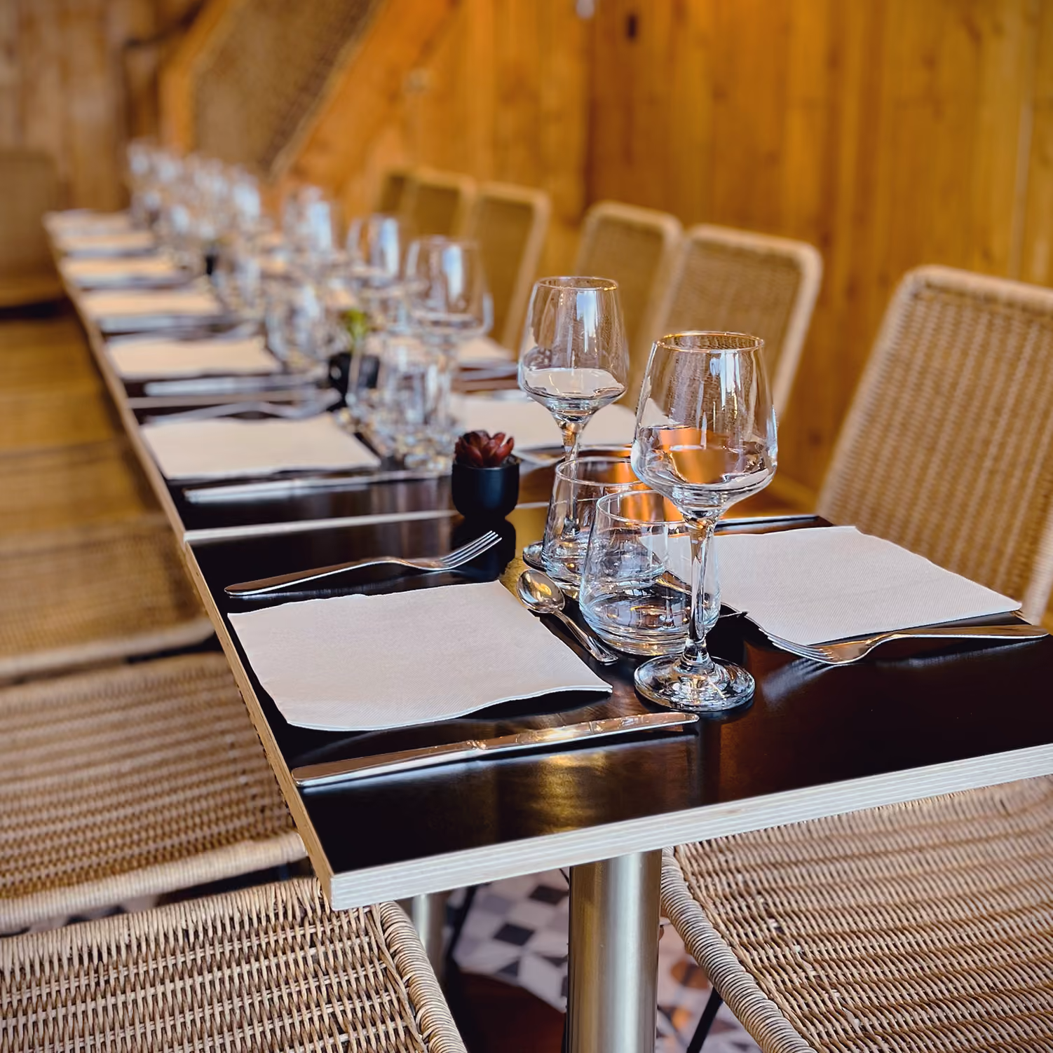 Grande table dressée du restaurant : verres à vin, couverts et serviettes, chaises en rotin et mur en bois