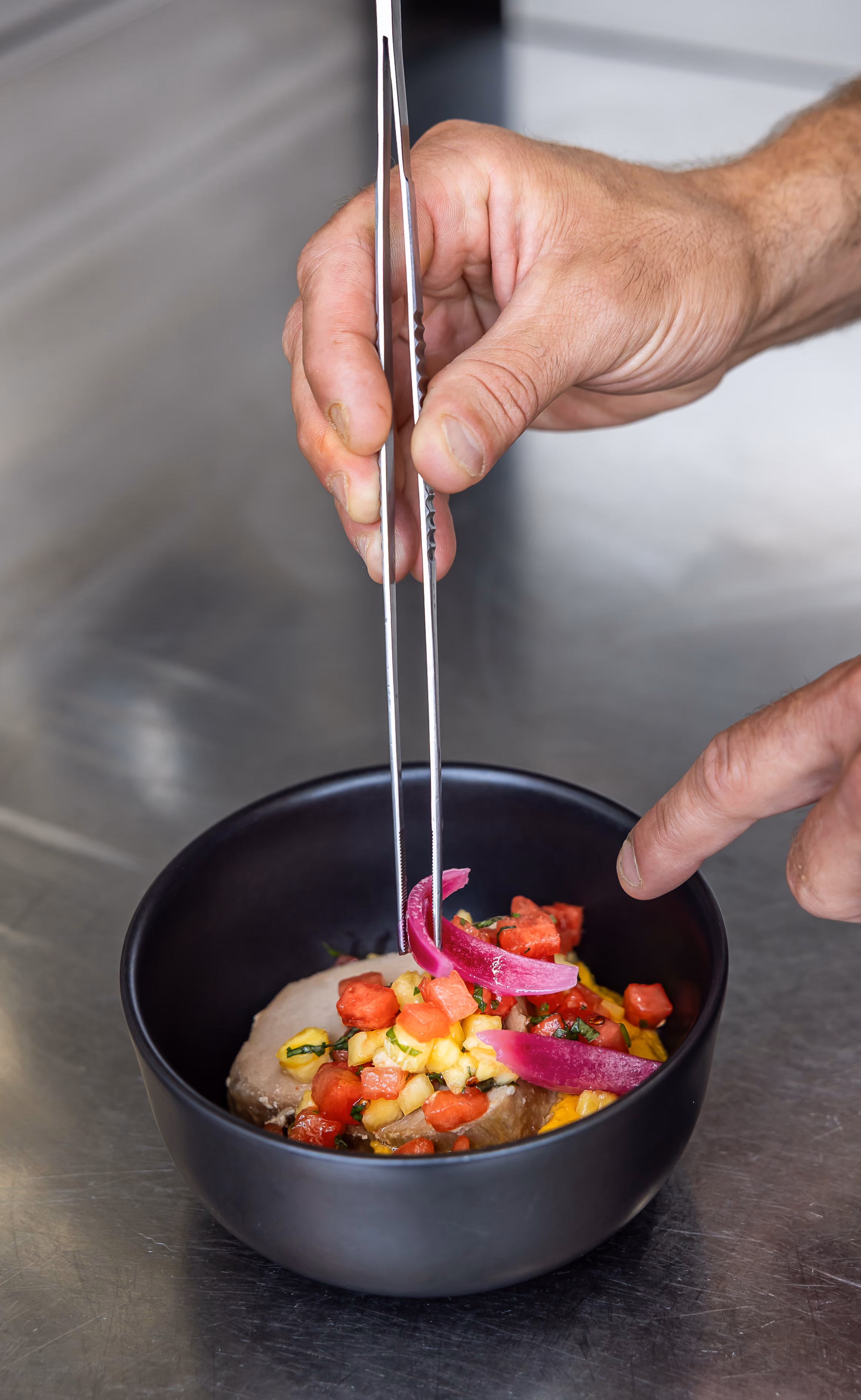 Chef déposant à la pince des oignons rouges marinés sur un rôti au tartare de pastèque, tomate et ananas dans un bol noir