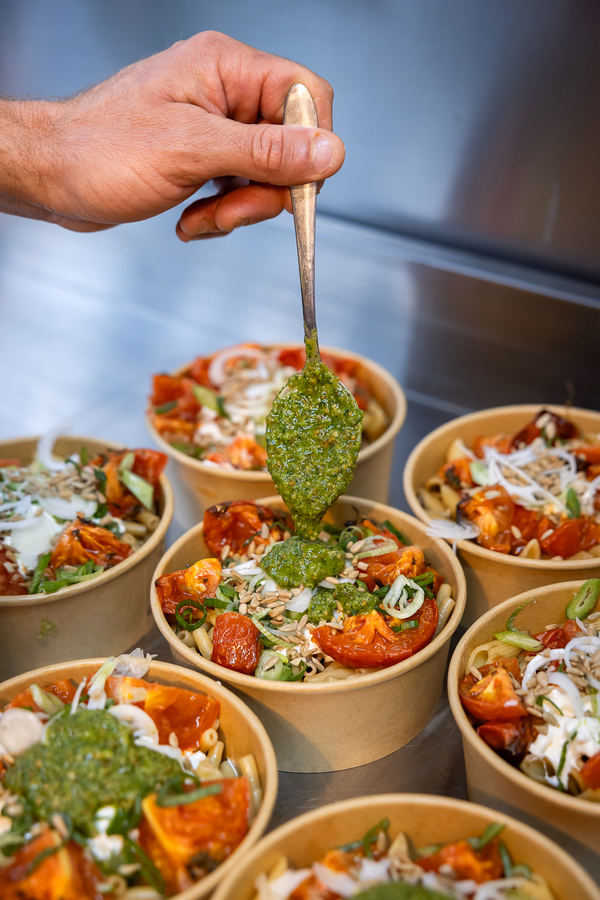 Main déposant une cuillère de pesto vert sur des bols de salade de pâtes aux tomates rôties, oignons et graines de tournesol en cuisine