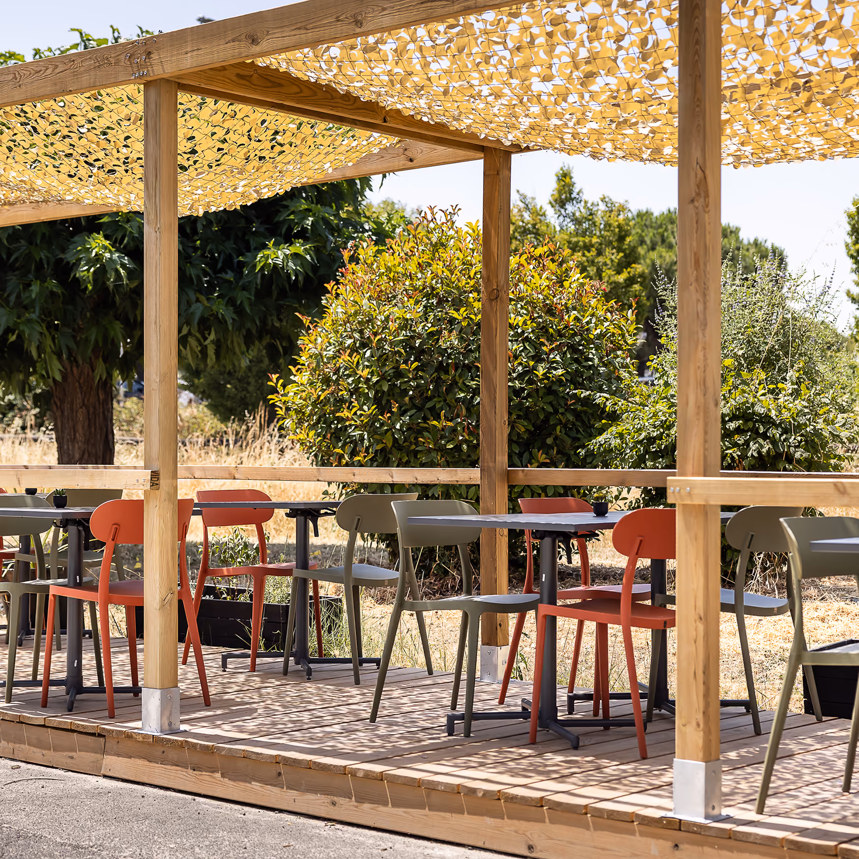 Terrasse en bois du restaurant sous pergola avec voile d'ombrage, chaises vertes et orange et cadre végétal
