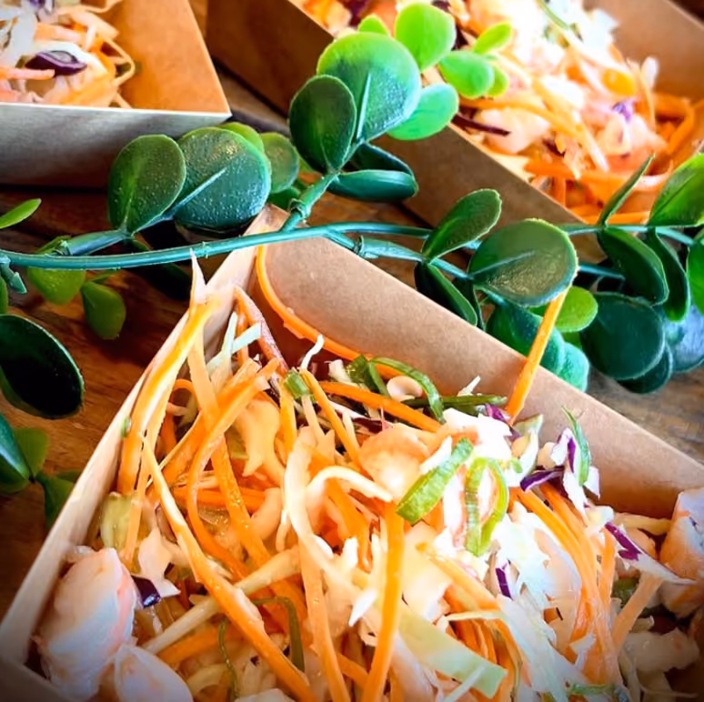 Salades de chou et carotte râpés aux crevettes en barquettes kraft, sur plan en bois avec verdure