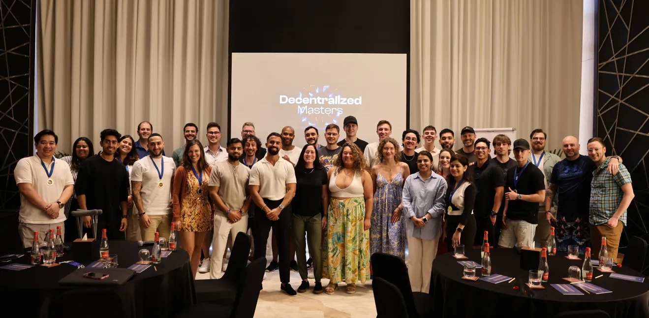 Group photo of 30 diverse adults standing in a conference room with tables and a screen that reads Decentralized Masters.
