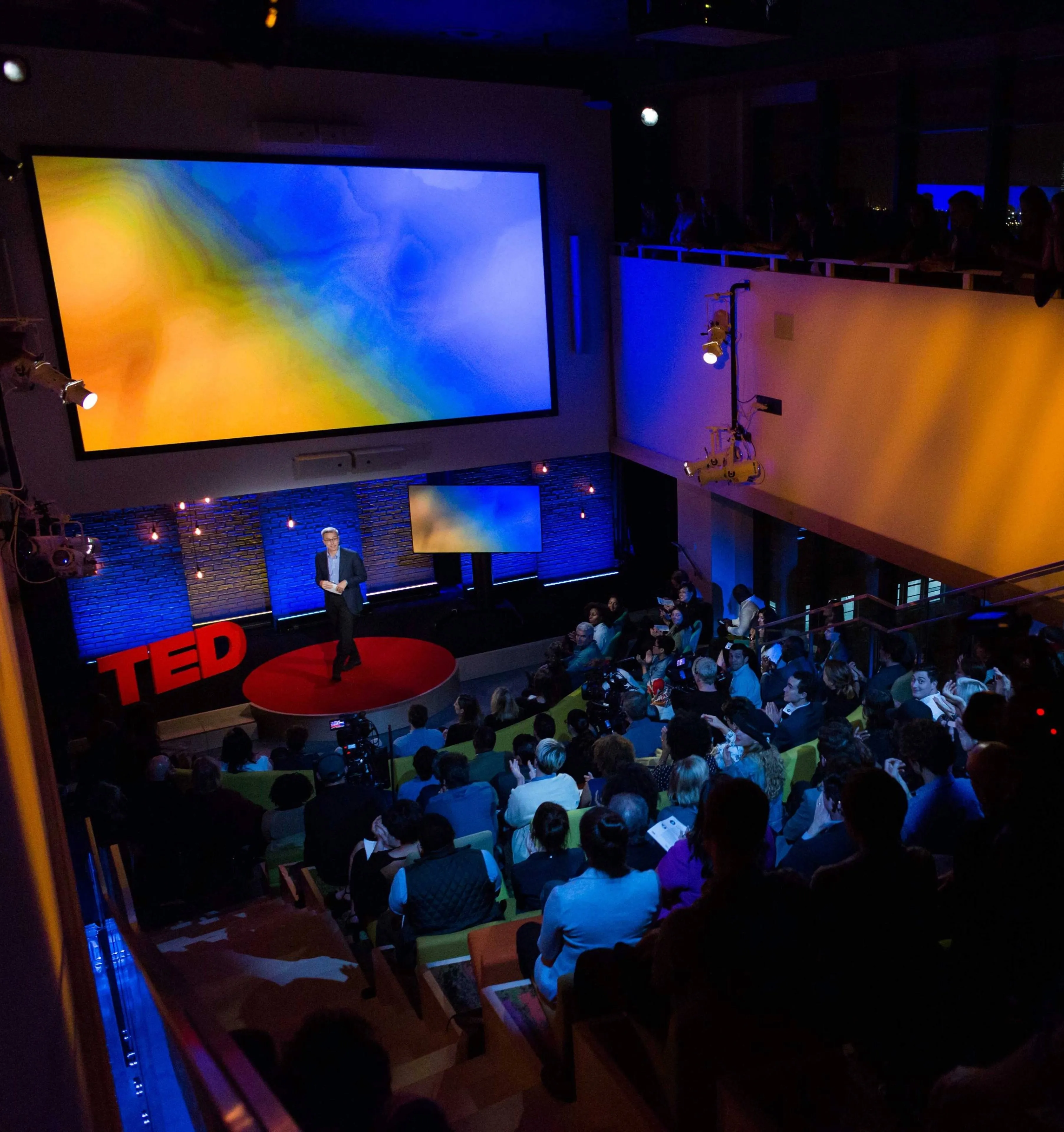A speaker on a red circular stage gives a TED talk in a dimly lit auditorium. A vibrant blue and yellow abstract image is displayed on a large screen behind them.