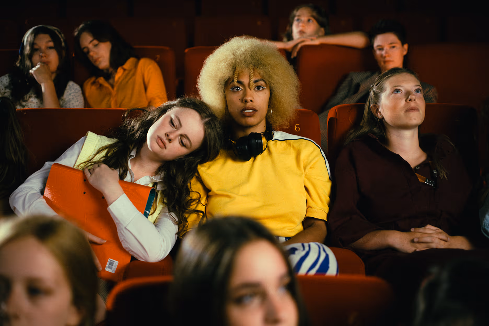 Young people sitting in red theater seats, with one person resting head on another's shoulder and another looking upward.