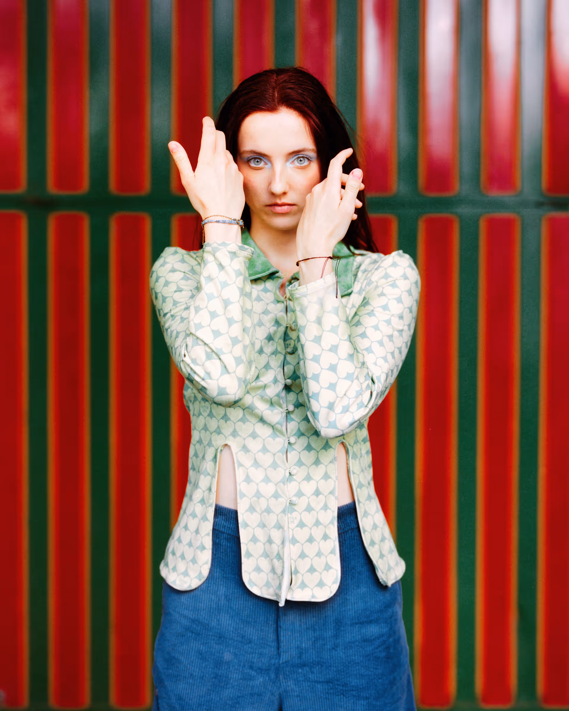 Woman with red hair in a heart-patterned green blouse and blue skirt posing with hands near face against green and red striped background.