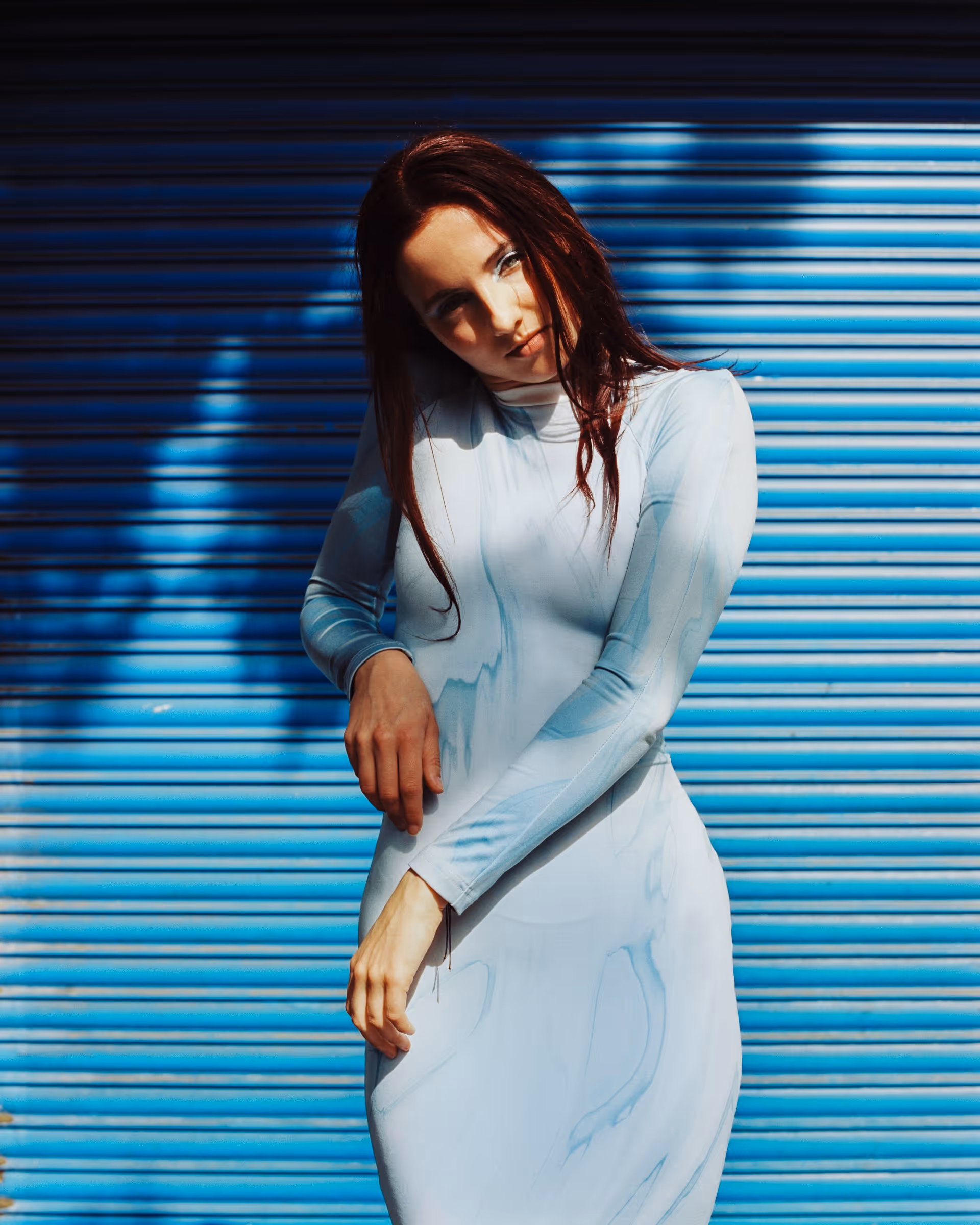 Young woman with long red hair wearing a light blue dress standing in front of a blue corrugated metal wall with shadows.