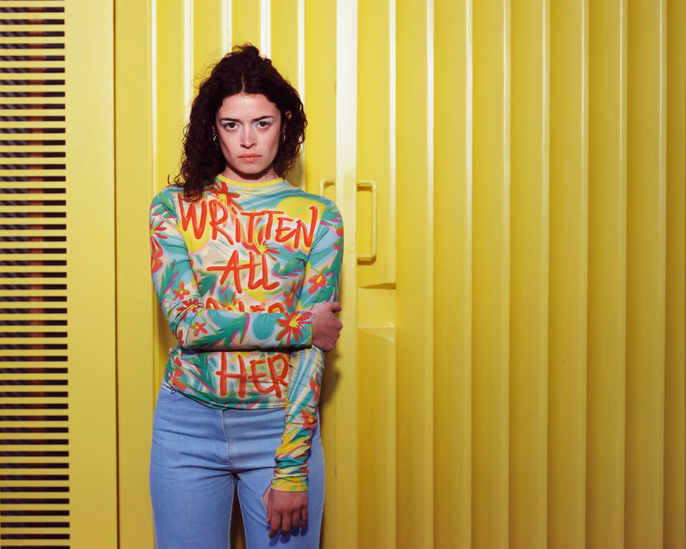 Woman with curly hair wearing a colorful floral long-sleeve shirt with 'WRITTEN ALL OVER HER' text, standing against a yellow corrugated metal door.