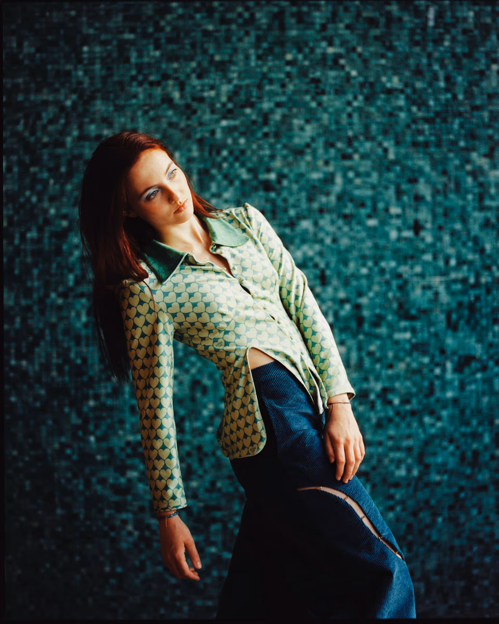Woman with long red hair wearing a green heart-patterned blouse and blue pants standing against a mosaic tile wall.