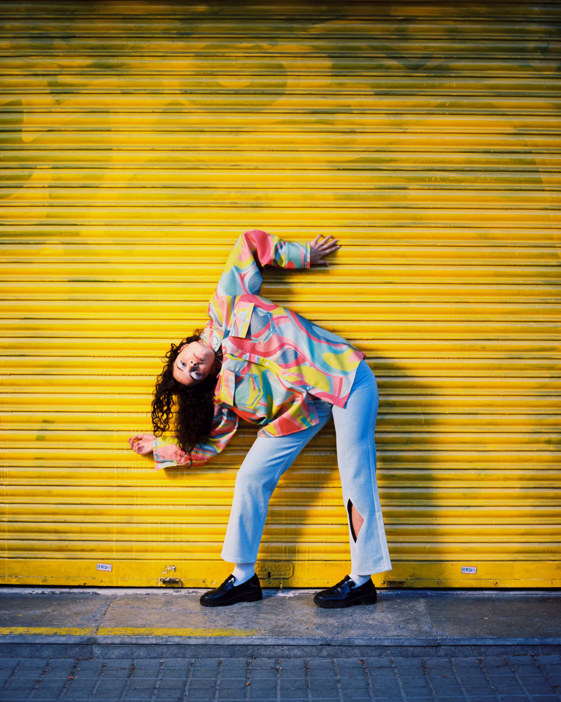 Person with curly hair wearing colorful patterned shirt and white pants posing leaning sideways against a yellow metal shutter.