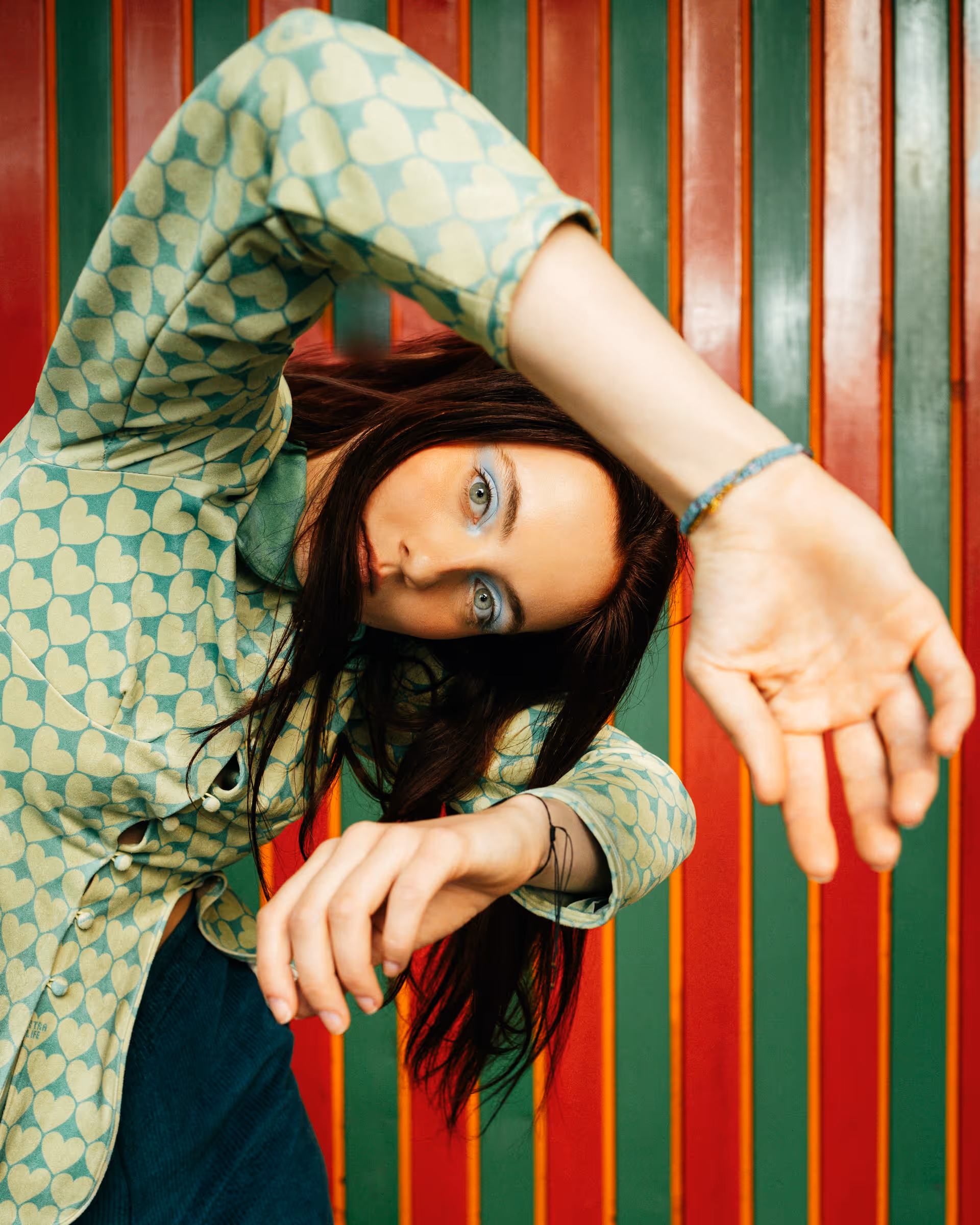 Woman with light blue eyeshadow and heart-patterned shirt posing with arms bent in front of a red and green striped background.