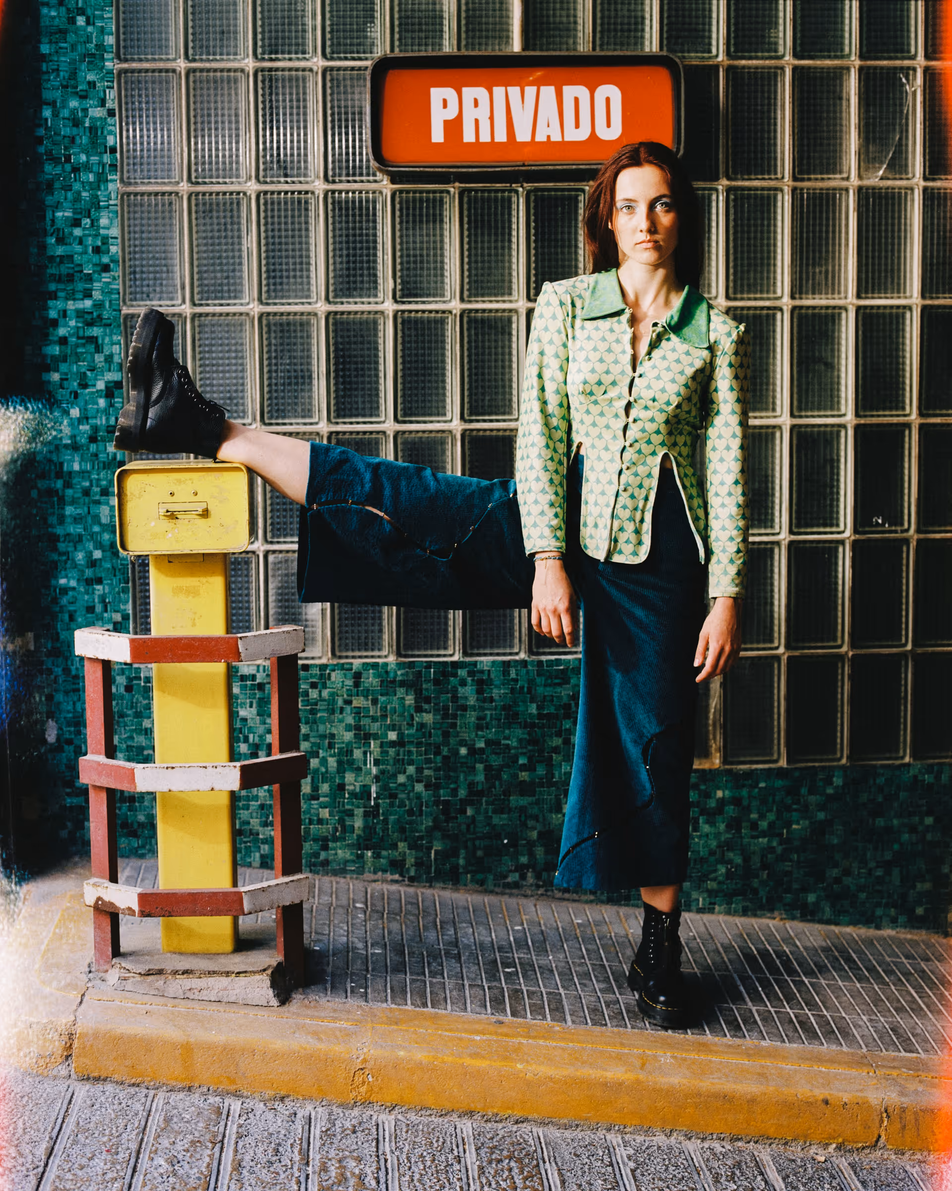 Woman in green patterned jacket and denim skirt stands with one leg resting on a yellow barrier under a sign that reads 'PRIVADO'.