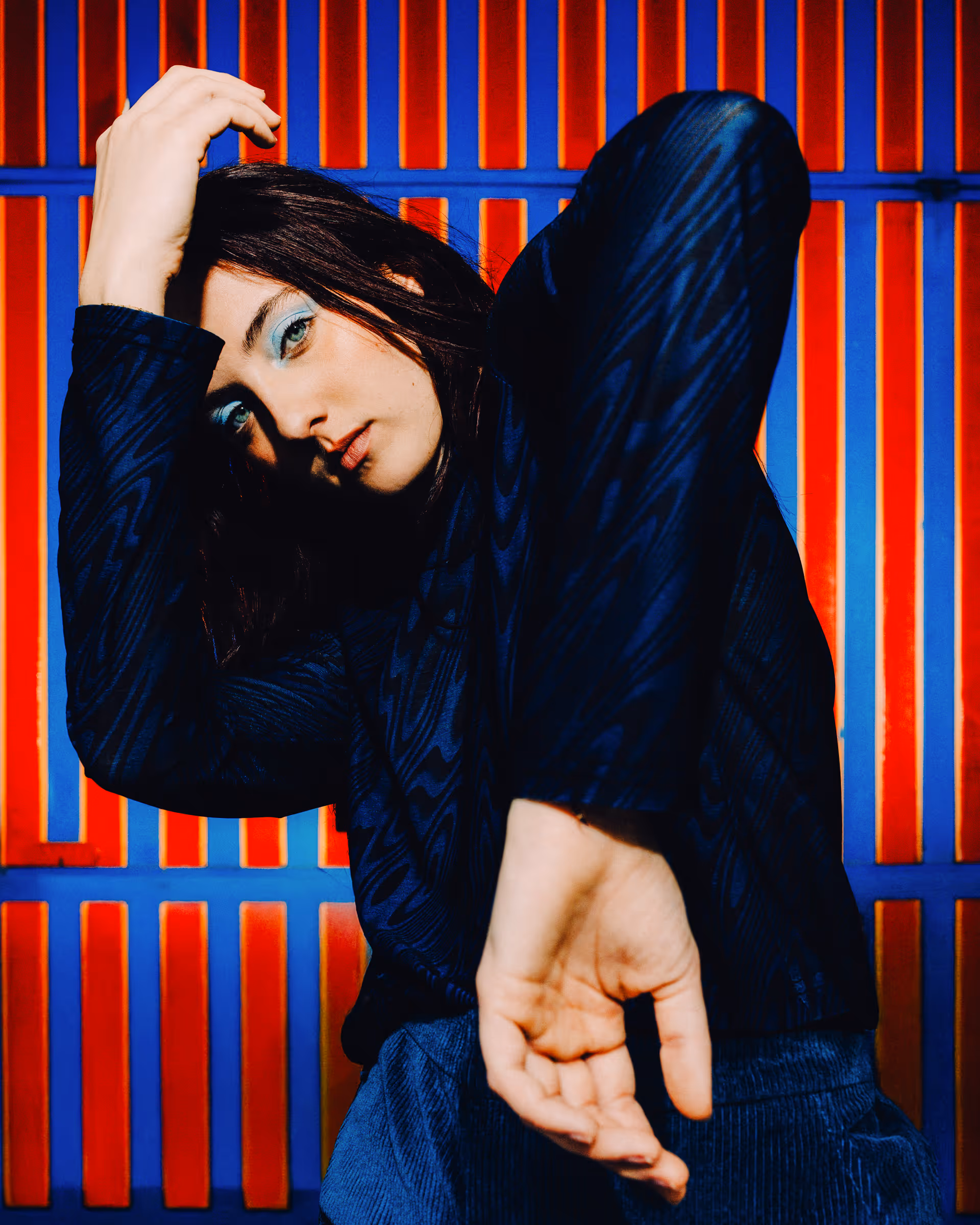 Woman posing with one arm raised and the other extended forward against a vibrant red and blue striped background.