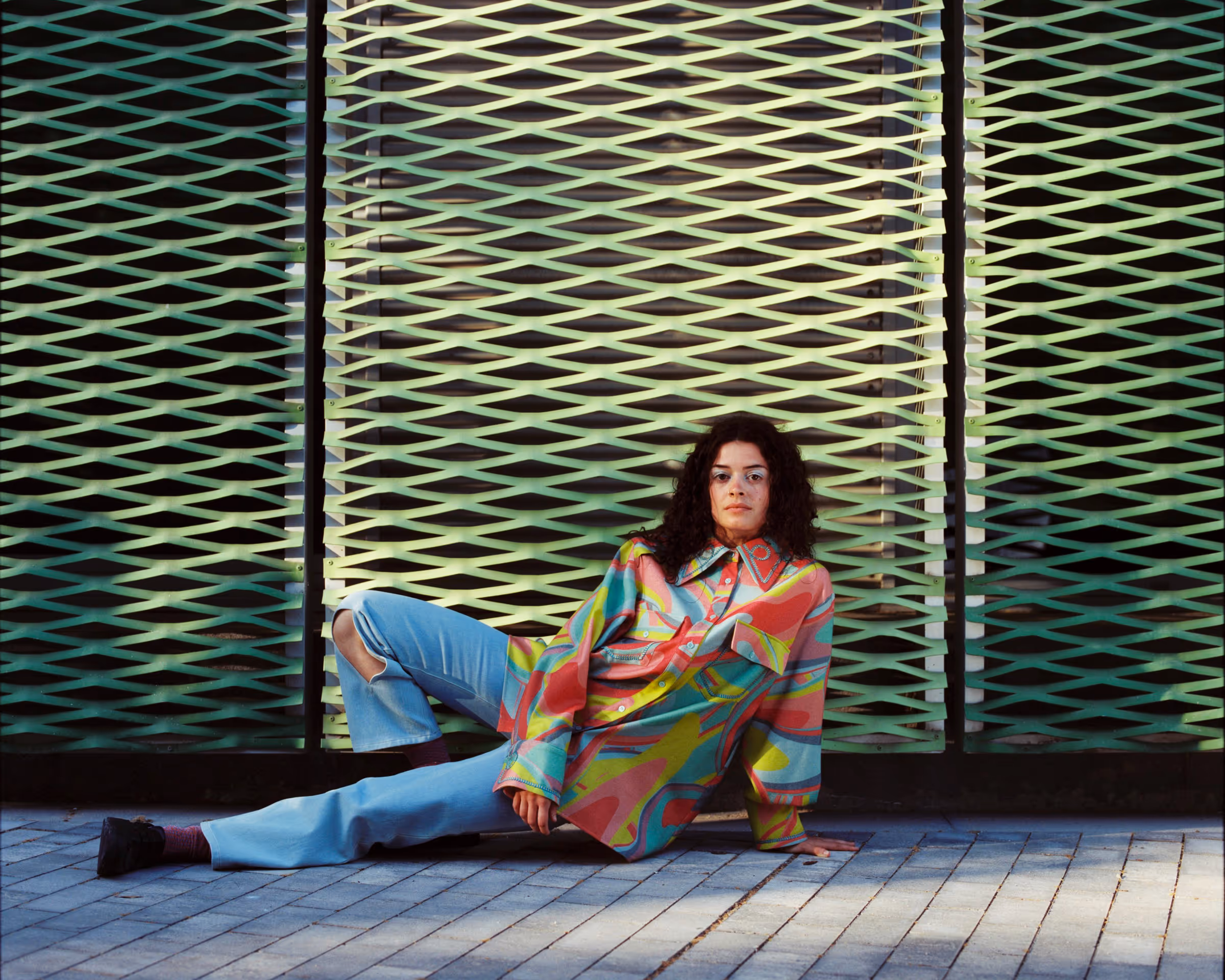 Woman with curly hair wearing a colorful oversized shirt and light blue ripped jeans sitting on the ground against a green patterned wall.