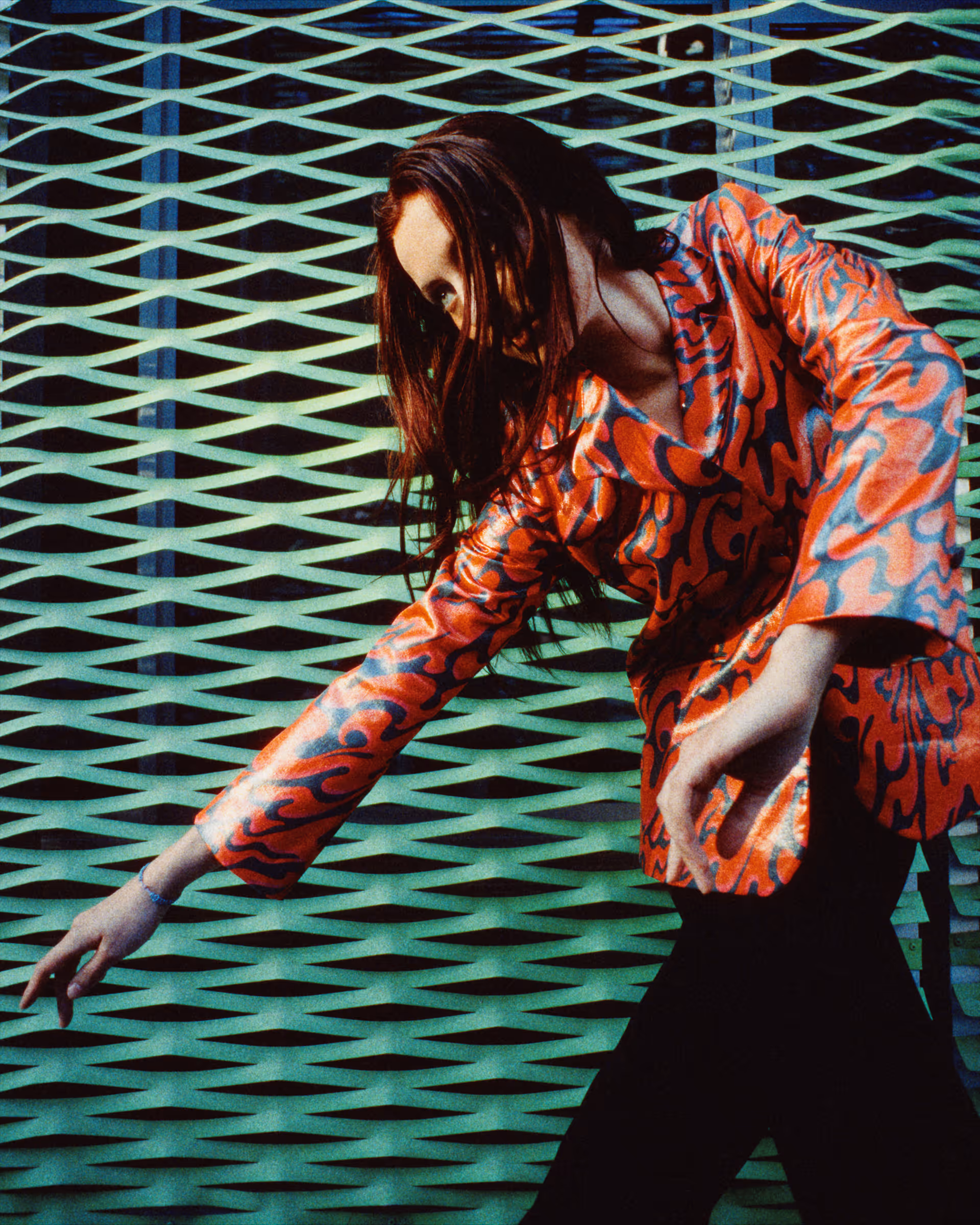 Woman with long brown hair wearing a shiny red-orange patterned jacket and black pants, posing with arms extended in front of a green patterned metal fence.