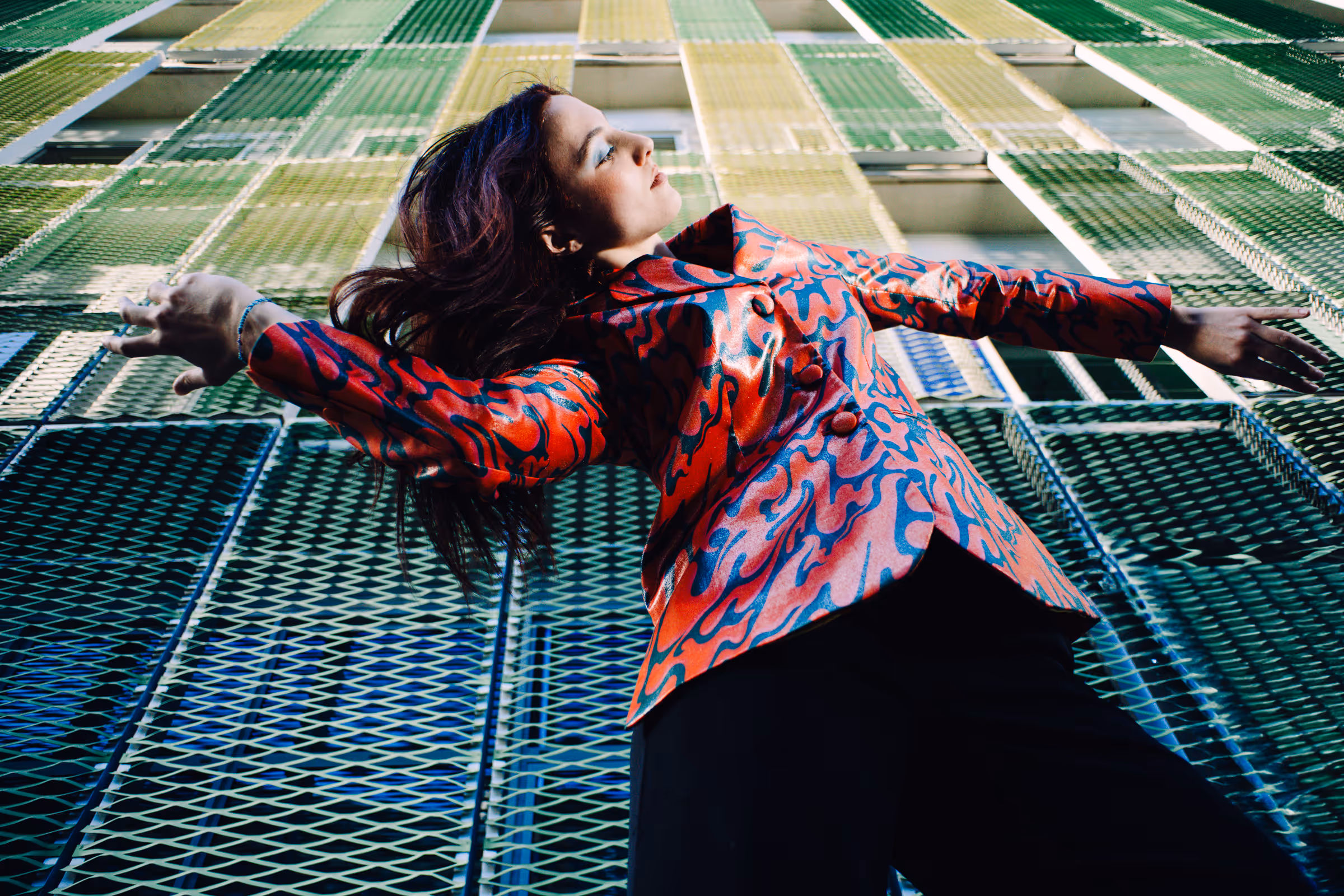 Woman wearing a red patterned jacket with arms outstretched in front of a green metal mesh building facade.